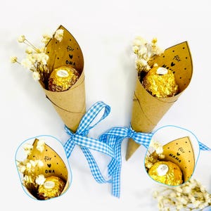 Könnte beinhalten: Zwei kleine Sträuße mit goldverpackten Schokoladenbonbons und getrockneten weißen Blumen. Die Sträuße sind in braune Papierkegel gewickelt und mit blau-weiß kariertem Band gebunden. Auf dem Papier stehen die Worte "für OMA" und "für MAMA".