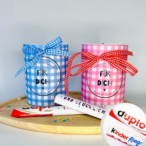 Könnte beinhalten: Zwei zylindrische Geschenkbehälter in blauem und rosa Vichy-Karo-Papier, jeweils mit einer passenden Schleife gebunden. Ein rundes Etikett auf jedem trägt die Aufschrift "FÜR DICH". Eine weiße Röhre mit "UND GLÜCKLICH" und zwei Schokoriegel.