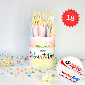 May include: A cylindrical gift container decorated with pastel colours and the text Alles Liebe zum Geburtstag, topped with paper candle cutouts. A red circular badge displays the number 18, and two chocolate bars are visible in the foreground.