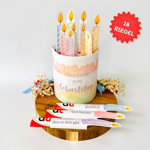 May include: A birthday-themed centerpiece featuring a cylindrical container decorated with pastel colors and paper candles. The container has the text "Alles Liebe zum Geburtstag". Several paper candles are placed inside the container, and there are three additional paper candles with text.
