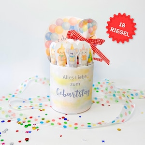 May include: A cylindrical gift box with a watercolour design, featuring the text "Alles Liebe zum Geburtstag." The box contains animal-shaped cutouts and is decorated with a red and white checkered ribbon and a balloon-themed topper. A red starburst label reads "18 RIEGEL."