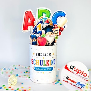 May include: A white cylindrical container decorated with colorful dots and the text ENDLICH SCHULKIND Schulstart 2026. It holds cutouts of the letters ABC, a globe, a rocket, a graduation gnome, a red apple, and a diploma.