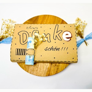 May include: A brown cardboard gift card with the German words "Danke schön!!!" in black script. The card features a hand-drawn design with a hand holding a rolled-up bill and is decorated with dried flowers.