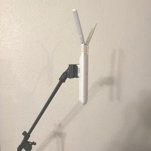 May include: A white wireless access point with two antennas, mounted on a black stand. The antennas are angled upwards. The stand is attached to a black tripod, set against a light beige wall.