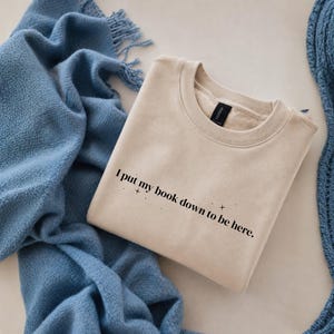 Könnte beinhalten: Ein beigefarbenes Sweatshirt mit Rundhalsausschnitt und dem schwarzen Text "I put my book down to be here.", ergänzt durch kleine Sternsymbole. Das Kleidungsstück hat einen gerippten Kragen, lange Ärmel und ein weiches Fleece-Innenfutter.
