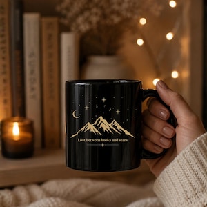 Könnte beinhalten: Schwarze Keramik-Tasse mit Berg- und Sternenmotiv. Die Tasse trägt den Text "Lost between books and stars" in Gold. Die Tasse wird von einer Person gehalten.