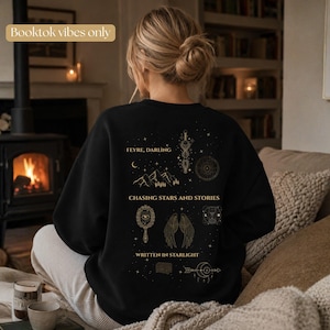 Könnte beinhalten: Schwarzes Sweatshirt mit goldfarbenen Grafiken und Text. Der Text lautet "Feyre, Darling", "Chasing Stars and Stories" und "Written in Starlight". Das Design zeigt Illustrationen mit Himmels- und Fantasy-Themen. Der Satz "Booktok vibes only" befindet sich oben links.
