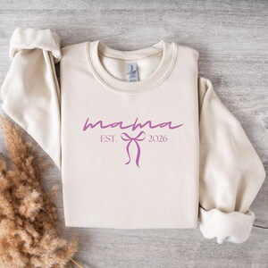 May include: Cream-colored sweatshirt with the word "mama" in a cursive font, along with "EST. 2026" and a bow graphic in a purple hue. The sweatshirt is folded, with the sleeves rolled up.