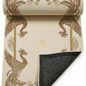 May include: Beige yoga mat featuring a brown phoenix design. The phoenix is framed by a gold border with cloud accents. The mat is partially rolled, revealing a black underside.