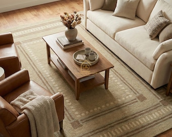 May include: A rectangular area rug featuring a geometric border pattern in shades of beige and brown. The rug is positioned under a wooden coffee table, two leather armchairs, and a cream-coloured sofa with several throw pillows in a living room.
