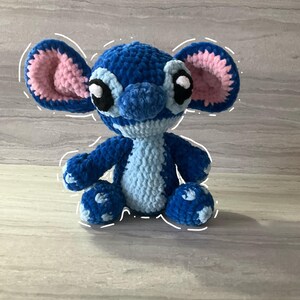 May include: A handmade blue and light blue crochet plush toy with large ears and black eyes. The plush toy is sitting on a light gray surface. The inside of the ears are pink.