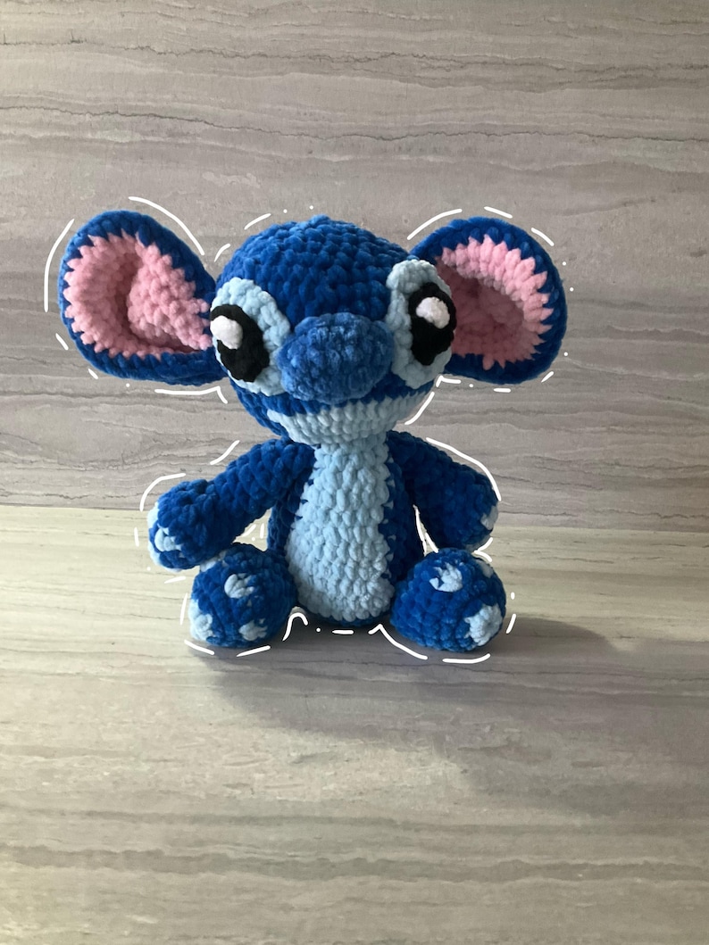 May include: A handmade blue and light blue crochet plush toy with large ears and black eyes. The plush toy is sitting on a light gray surface. The inside of the ears are pink.