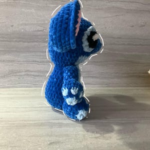 May include: A handmade blue crochet Stitch doll, featuring large ears with pink inner details, a round body, and small arms and legs. The doll has a black and white eye and a small nose. The plush toy is made of soft, textured yarn.