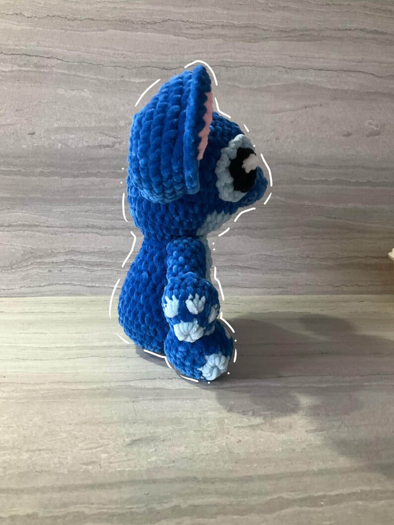 May include: A handmade blue crochet Stitch doll, featuring large ears with pink inner details, a round body, and small arms and legs. The doll has a black and white eye and a small nose. The plush toy is made of soft, textured yarn.
