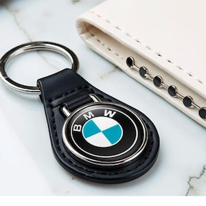 May include: Black leather keychain with a silver ring and a circular emblem featuring the BMW logo in black, white, and teal. The keychain is next to a white leather wallet.