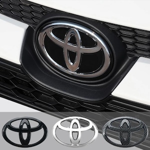 Puede incluir: Primer plano del emblema de Toyota en la parrilla delantera de un coche. El emblema es plateado y está sobre un fondo negro. Se muestran emblemas adicionales en negro, plateado y fibra de carbono.