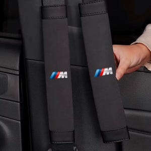 May include: Black seat belt shoulder pads featuring the "M" logo in blue, red, and white. These pads are made of a soft material and are designed for comfort and protection. The pads are shown in a car interior.
