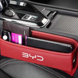 May include: Red car seat organizer with the white text "BYD". The organizer is filled with a wallet, keys, credit card, and a smartphone. The organizer is made of a red material and is placed between the car seats.