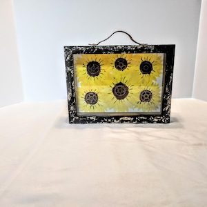 May include: A rectangular black framed art piece featuring a sunflower design. The artwork has a distressed finish and a brown handle. The sunflowers are yellow with brown centers, set against a yellow background.