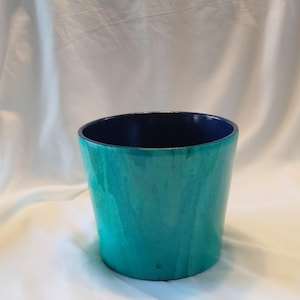 May include: A teal-colored ceramic planter with a dark blue interior. The planter has a slightly flared shape and a glossy finish. The background is a white, draped fabric.
