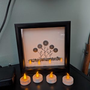 以下が含まれることがあります： 黒い正方形のシャドーボックスフレーム。白い背景に、4本の花の茎と、その先端にボタンをあしらった手描きのインク画が描かれています。茎の根元には小さな小石が並べられています。フレームの前にはオレンジ色の炎が灯る電池式のティーライトキャンドルが4つ置かれ、フレームの中にもさらに4つ置かれています。