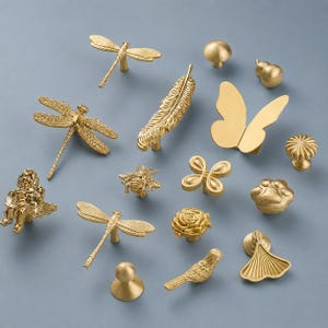 May include: A collection of gold-colored cabinet knobs and pulls in various designs, including dragonflies, a feather, a butterfly, a bee, a flower, a bird, and a paw print. The hardware is displayed on a light blue background.