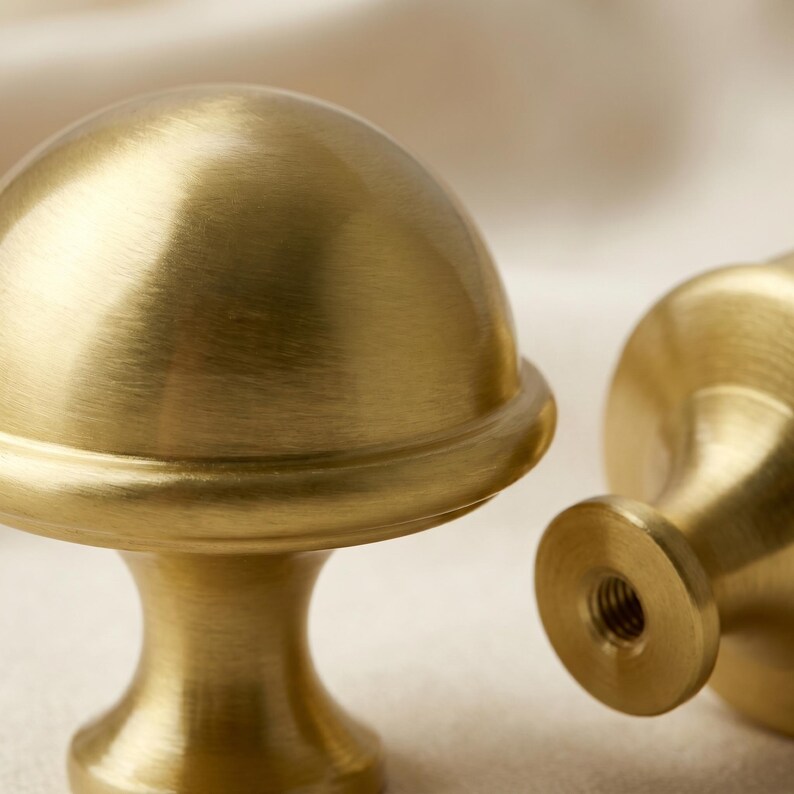 Op de afbeelding: Twee messing kastknoppen met een paddenstoelvormig ontwerp. De knoppen hebben een geborstelde afwerking en een schroefdraadbasis voor eenvoudige installatie. De knoppen hebben een warme goudkleur en zijn ideaal om een vleugje elegantie toe te voegen.