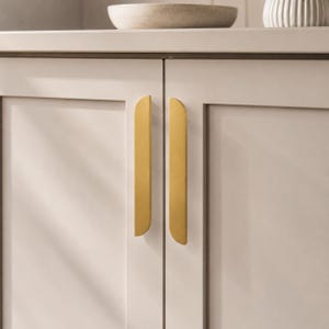 May include: Close-up of a cabinet with two doors and gold-colored, vertical bar handles. The cabinet doors are a light neutral color, and the handles have rounded edges. A shadow falls across the left side of the cabinet.