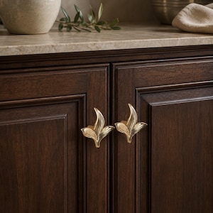 May include: Close-up of two gold-colored leaf-shaped cabinet pulls mounted on a dark brown wooden cabinet door. The pulls are symmetrical and have a detailed, textured design. The cabinet doors have a recessed panel design.