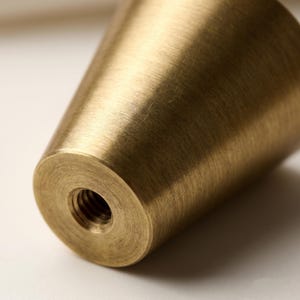 Puede incluir: Primer plano de un tirador de armario de lat&oacute;n macizo con forma c&oacute;nica. El tirador tiene una base roscada para una f&aacute;cil instalaci&oacute;n y un acabado cepillado. El lat&oacute;n tiene un tono dorado c&aacute;lido.