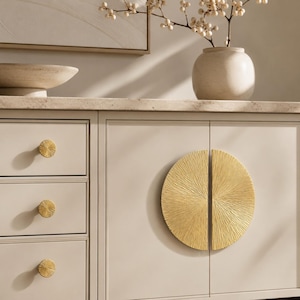 May include: A light beige cabinet with gold hardware. The cabinet has three drawers with round, textured gold knobs. The cabinet doors feature a large, textured gold circular handle. A decorative bowl and vase with flowers sit on top.