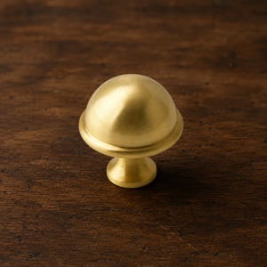 Op de afbeelding: Een messing kastknop met een afgeronde bovenkant en een cirkelvormige basis. De knop heeft een gladde, gepolijste afwerking en is ontworpen voor meubels. De hardware heeft een warme goudkleur en is geplaatst tegen een donkere houten achtergrond.