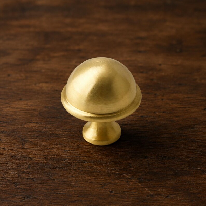 Op de afbeelding: Een messing kastknop met een afgeronde bovenkant en een cirkelvormige basis. De knop heeft een gladde, gepolijste afwerking en is ontworpen voor meubels. De hardware heeft een warme goudkleur en is geplaatst tegen een donkere houten achtergrond.