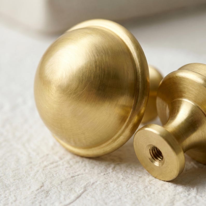 Op de afbeelding: Close-up van verschillende messing kastknoppen met een geborstelde afwerking. De knoppen hebben een afgeronde, koepelachtige vorm en een schroefdraadbasis voor eenvoudige installatie. De warme goudkleur voegt een vleugje elegantie toe.