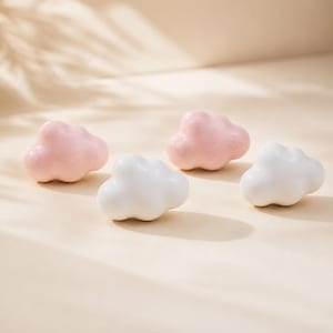 May include: Four cloud-shaped decorative objects. Two are white, and two are pink. The objects are arranged on a light beige surface, with shadows suggesting a soft, natural light.