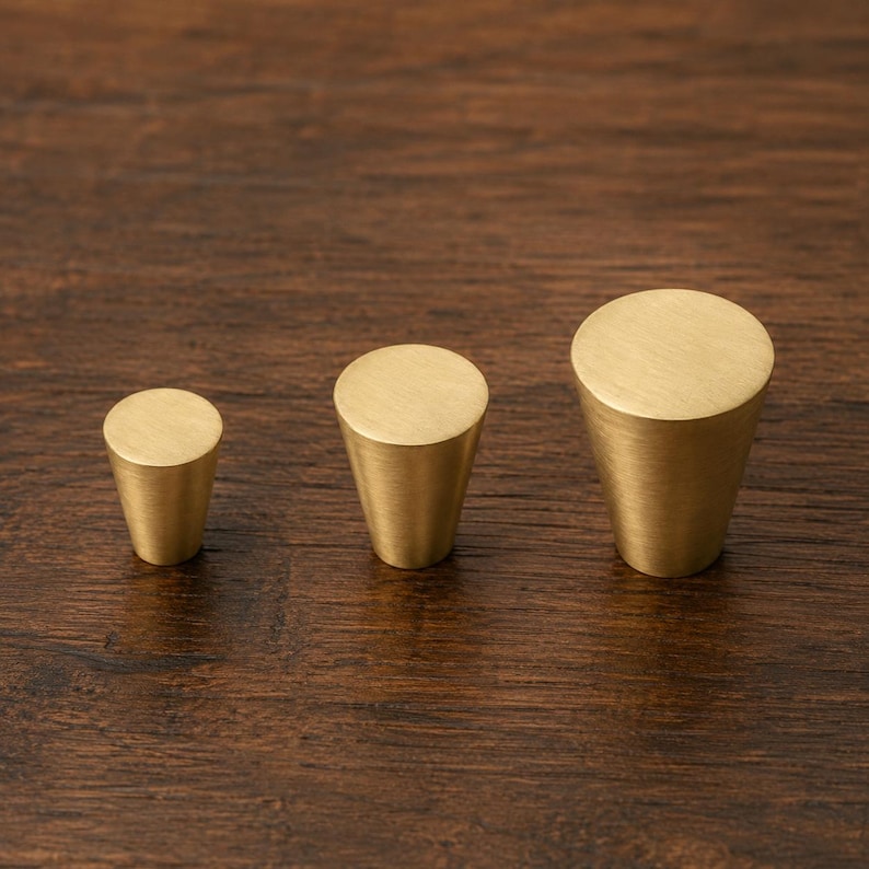 Puede incluir: Tres pomos de lat&oacute;n para armarios de diferentes tama&ntilde;os est&aacute;n dispuestos sobre una superficie de madera oscura. Cada pomo tiene forma c&oacute;nica con una parte superior plana y circular. Los pomos son de metal brillante de color dorado.