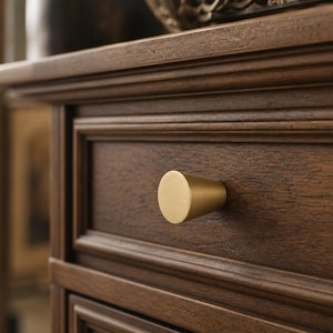 Puede incluir: Primer plano de un caj&oacute;n de madera marr&oacute;n oscuro con un pomo c&oacute;nico de color dorado. El caj&oacute;n tiene un dise&ntilde;o de panel empotrado y forma parte de un mueble m&aacute;s grande. La veta de la madera es visible.