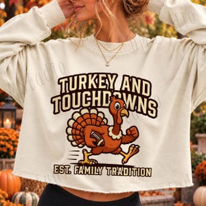 Pode incluir: Uma camisola curta de cor creme com um gráfico de um peru castanho a correr enquanto segura uma bola de futebol americano. O texto TURKEY AND TOUCHDOWNS está acima do peru e EST. FAMILY TRADITION abaixo, numa fonte castanha escura.