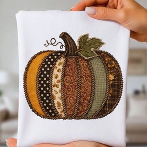 May include: A white fabric item featuring a central pumpkin graphic composed of vertical patchwork segments. The segments display varied patterns including yellow, black with white dots, cream with acorns, brown paisley, green, and brown plaid.