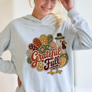 Op de afbeelding: Een lichtgrijze hoodie met een afbeelding van een kalkoen met een staart in patchworkstijl in tinten oranje, groen en bruin. De kalkoen draagt een pelgrimshoed en de tekst "Grateful & Full" is in een vetgedrukt, oranje cursief lettertype gedrukt.