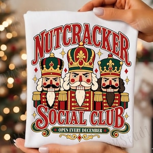 May include: A white t-shirt featuring a graphic of three nutcracker figures in red, green, and gold uniforms with crowns. The text reads NUTCRACKER SOCIAL CLUB and OPEN EVERY DECEMBER in red and green lettering with small gold star accents.