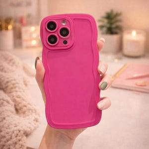 May include: A bright pink phone case with a wavy edge design. The case has a raised border around the camera lenses. The phone case is held in a person's hand.