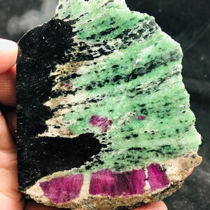 May include: A flat, polished slab of zoisite mineral featuring a vibrant green base with prominent black hornblende inclusions and several distinct, deep magenta-colored ruby crystals embedded along the lower edge of the stone specimen.