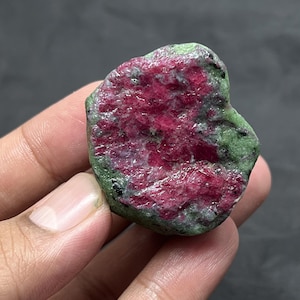 May include: A raw ruby gemstone, displaying a vibrant red core surrounded by a green exterior. The natural stone has an uneven texture and shape, highlighting its unrefined form. The ruby is held in a hand against a dark backdrop.