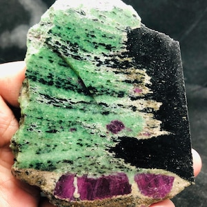 Pode incluir: Um espécime mineral bruto com uma base verde vibrante e estrias e inclusões pretas proeminentes. A pedra exibe várias secções cristalinas de cor magenta profunda ao longo da borda inferior e espalhadas por toda a superfície.