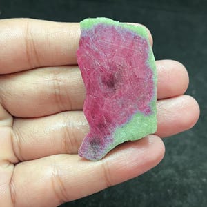May include: A flat, irregularly shaped slice of ruby zoisite mineral featuring a vibrant magenta-pink center surrounded by a pale lime-green border. The surface displays a natural crystalline texture with visible geometric patterns and inclusions.