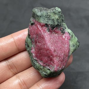 May include: A raw gemstone with a striking pink core and a green and black outer layer. The stone's surface is unpolished, revealing its natural texture. The gemstone is held in a hand, showing its size and unique characteristics.