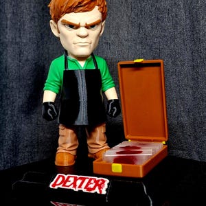May include: A stylised figurine of a character with ginger hair, wearing a green shirt, black apron, and black gloves. Beside the figure is an open brown case containing glass slides with red markings. The base features the text DEXTER in white.