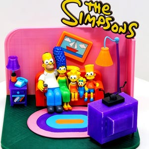 Pode incluir: Um diorama impresso em 3D da sala de estar dos Simpsons com Homer, Marge, Lisa, Maggie e Bart sentados num sofá laranja. A cena inclui uma televisão roxa, uma parede rosa com um quadro de um veleiro, uma mesa lateral roxa com um telefone azul e um tapete.
