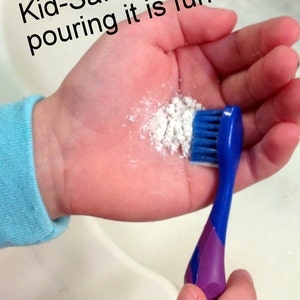 May include: A child's hand holding a blue and purple toothbrush with white powder and the text "Kid-Safe! Plus, pouring it is fun!" The toothbrush has blue bristles and a purple handle.